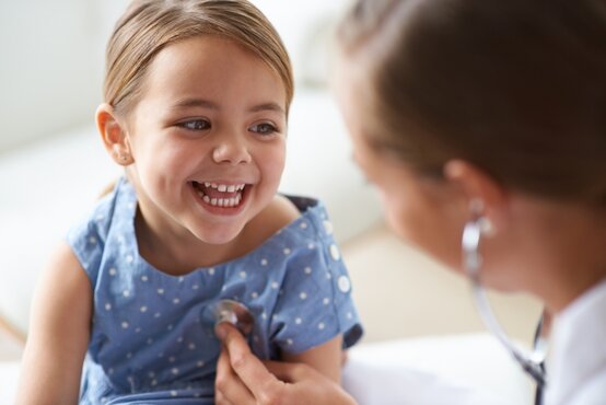 Young girl in blue dress laughing, doctor holding stethoscope to her chest, bright blurred background