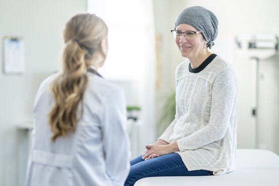 Woman with grey headscarf and glasses sitting on examination couch, smiling and talking to doctor in white coat in bright room