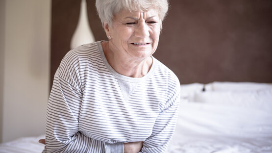 An elderly person with grey hair is sitting on a bed, holding their stomach with both hands, their face contorted with pain.
