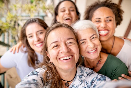 Five women of different ages smile and hug each other tightly for a selfie