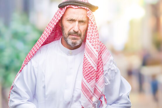 Man of middle age in white thobe and red and white keffiyeh with a serious expression on his face outside