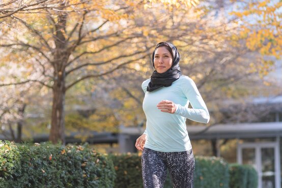 Woman in a black hijab, blue long-sleeved shirt, and patterned leggings jogs in a sunny autumn park