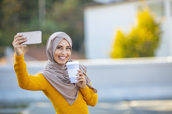 Smiling woman in a light brown hijab and yellow sweater takes a selfie outdoors, holding a smartphone and takeaway coffee cup